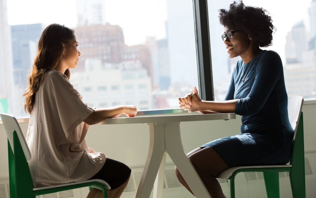 Deux femmes en pleine discussion assises à une table dans un environnement urbain lumineux, représentant un moment d'échange et de soutien mutuel.